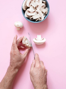 男性手用小刀切片新鲜美味的蘑菇 Champignons 在一个盘子和粉红色的背景。垂直