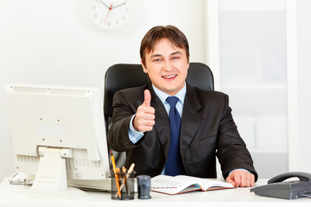 微笑着坐在办公室桌前和显示 ges 竖起大拇指的现代商人