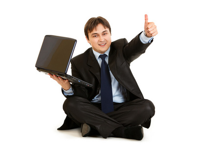 微笑着坐在地板上用笔记本电脑显示手势竖起大拇指的商人