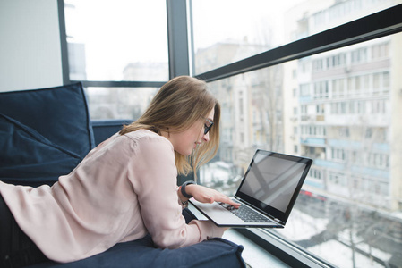 女孩自由职业者工作的笔记本电脑, 而躺在沙发附近的窗口。办公室免费工作