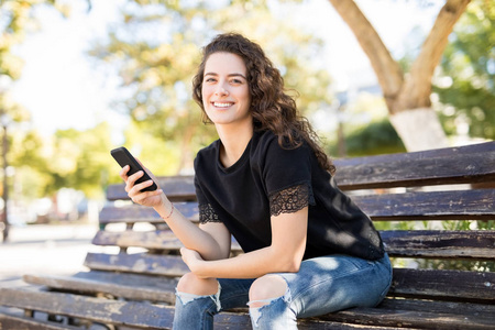 时尚的年轻女子坐在公园长凳上的智能手机