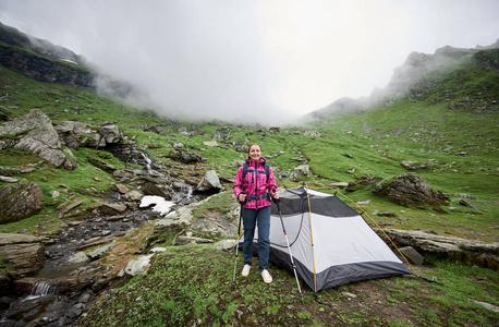 在罗马尼亚山脉山谷的帐篷附近, 有背包和徒步旅行的女性游客。在背景下, 山上有雾。