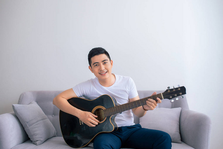 英俊的亚洲男子玩吉他坐在沙发上在家