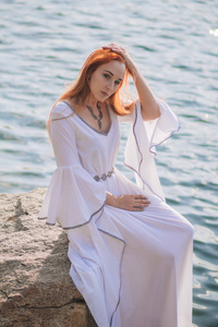 美丽的红头发的女孩漂浮在水中。洛基海岸。翡翠水中的一种白色古董礼服模型。中世纪的形象。幻想