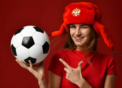 俄罗斯风格风扇运动女子运动员在红色制服和耳朵襟翼帽子举行足球庆祝愉快的微笑