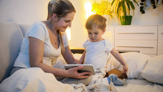 美丽的年轻女子睡衣与她的2岁的儿子看漫画在数字平板电脑晚上