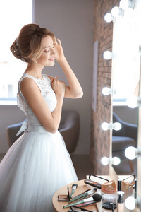 美丽的年轻新娘在白色婚纱礼服与化妆师附近的专业艺术家在室内镜子