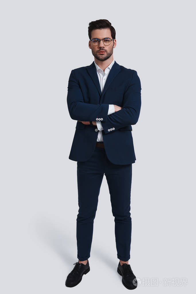 自信的年轻人在充分的西装看着相机和保持双臂交叉, 而站立反对灰色背