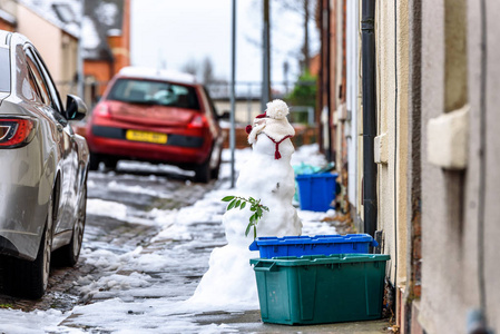 在典型的英国道路小径上的自制雪人的多云的冬天天的看法在空的回收箱旁边