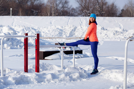 年轻妇女在明亮的蓝色帽子, 橙色毛衣和麋鹿微笑和舒展在奔跑在一个运动场在一个明亮的冬天天