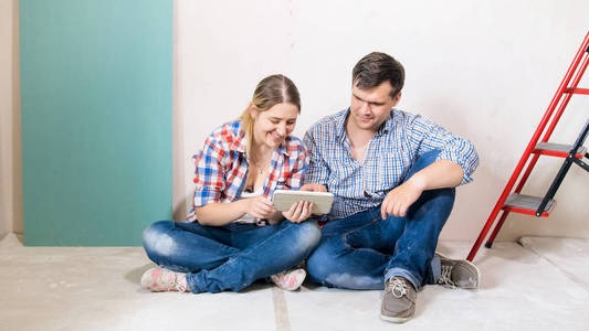 微笑的年轻夫妇坐在地板上的新房子下建设和浏览互联网