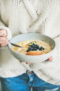 健康的冬季早餐在床上。穿羊毛毛衣和牛仔裤的女人吃纯素杏仁牛奶燕麦粥, 浆果, 水果和杏仁