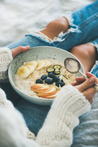 健康的冬季早餐在床上。妇女在羊毛毛衣和破旧的牛仔裤吃纯素杏仁牛奶燕麦粥与浆果