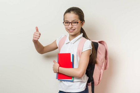 女孩学校学生, 用背包和笔记本显示竖起大拇指标志, 背景明亮的墙壁在学校