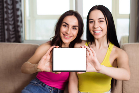两个微笑的美丽的现代女朋友坐在沙发上拿着白色的片剂在他们的手中, 并显示它的相机