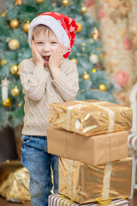 一个戴红帽的男孩坐在圣诞树旁, 带着礼物