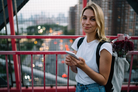 青年女游客背着背包带花的半长肖像城市用手机导航, 时髦女孩站在城市桥梁与 smartphon