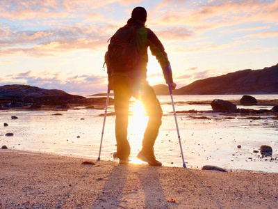 快乐的残疾人在海滩上拿着他的前臂拐杖头顶, 温暖的日落