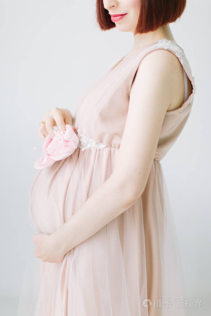 幸福的孕妇站立抱着粉红色的鞋子为婴孩庄稼的外形视图