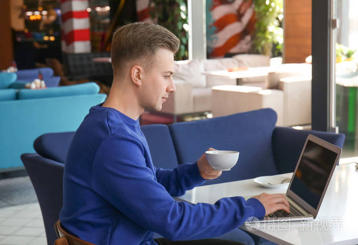 年轻的自由职业者喝咖啡, 而在桌子上的笔记本电脑在咖啡馆照片-正版