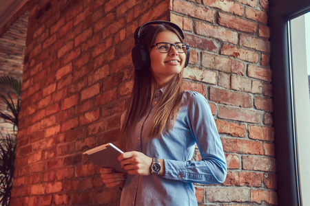 戴眼镜和蓝色衬衣的年轻黑发女郎。微笑着站在砖墙上, 用平板电脑听音乐
