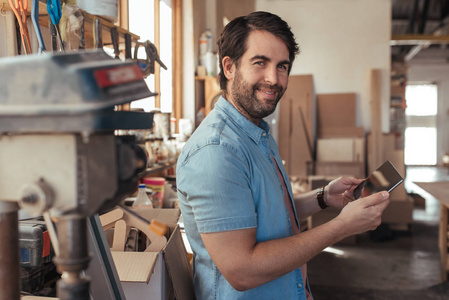 微笑的年轻木工的肖像站在工作台上的工具在他的车间与数字平板电脑在线工作