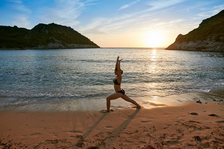 剪影瑜伽练习在日落。在美丽的海滩上做瑜伽练习的年轻女子。科孚, 希腊