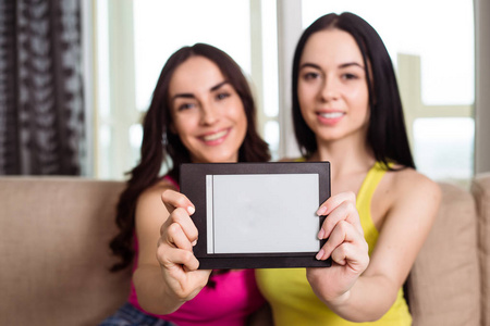 两个微笑的美丽的现代女朋友坐在沙发上拿着白色的片剂在他们的手中, 并显示它的相机
