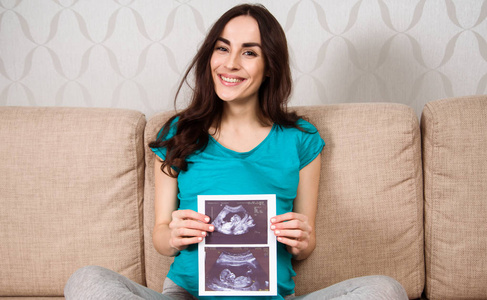 微笑着美丽的孕妇坐在沙发上, 拿着一张超声波照片, 并把它展示给相机。等待孩子