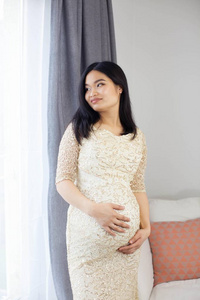 美丽的年轻亚洲孕妇站在窗口和触摸她的肚子。怀孕.健康.母亲.一个孕妇的舒适肖像。舒适