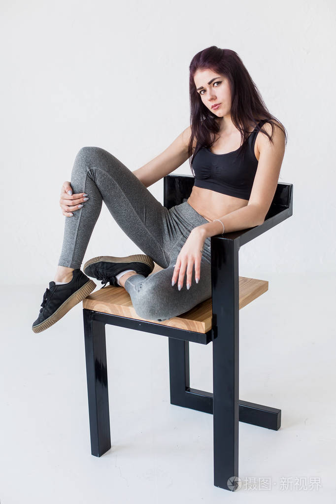 在白色演播室坐在椅子上的年轻迷人的体育妇女的全长画像 sportsuit