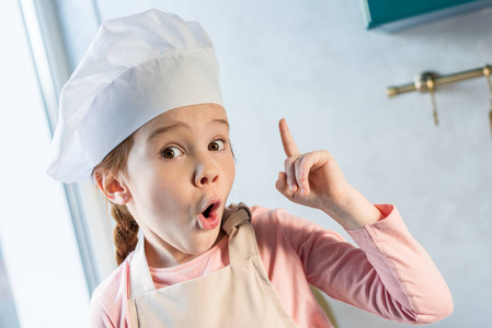 可爱的孩子在厨师帽子指着手指和看在厨房的摄像头