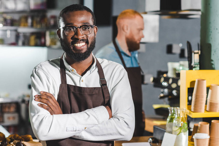 年轻的微笑非洲裔美国男性咖啡师在围裙与同事站在后面的肖像