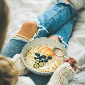 健康的冬季早餐在床上。穿着羊毛衫和破旧牛仔裤的女人吃素食杏仁牛奶燕麦粥, 浆果, 水果和杏仁