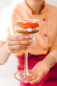 特写的女性手捧着高高的玻璃蛋糕站与艺术装饰甜点制成的海绵层, 奶油, 装饰花和白色巧克力戒指