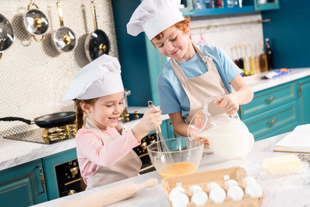 愉快的小孩子在厨师帽子和围裙一起准备面团在厨房