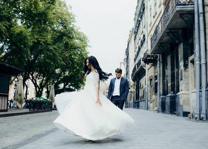 在新郎的模糊背景下, 穿着漂亮笑容的黑发新娘在长婚纱中旋转。城市街道位置