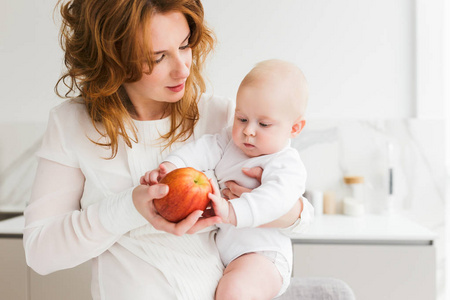 美丽的母亲的肖像站在厨房, 抱着她可爱的小宝宝和大红苹果在手隔绝