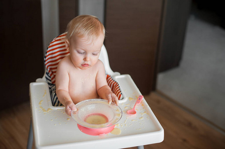 小可爱的女孩坐在高高的椅子和学习用勺子自己吃。有趣的快乐婴儿涂抹在桌子上的食物
