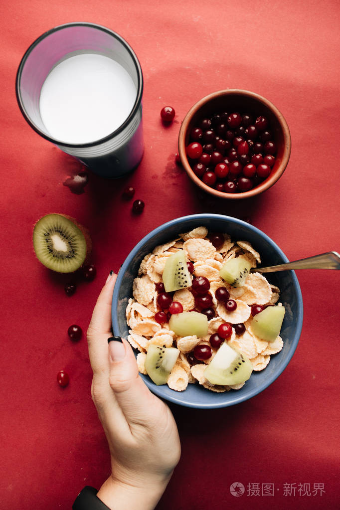 女性手捧碗, 配以猕猴桃片和蔓越莓, 健康早餐的红色背景美味酥皮玉米