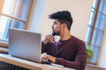 英俊的美国黑人男子在休闲服装拿着一杯咖啡和使用笔记本电脑