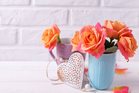 鲜艳的玫瑰在蓝色的杯子和装饰的心脏白色木质背景反对白色墙壁。花静物。选择性对焦。文本位置