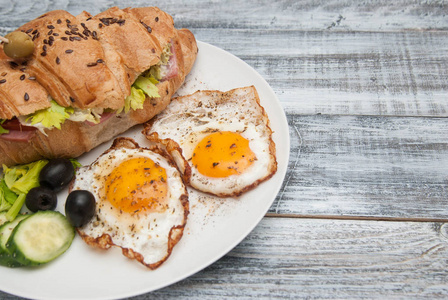 白盘子配煎蛋和羊角面包三明治, 配黄瓜, Tomatoe 和橄榄。早餐.灰色的木质背景。仿古风格与复制粘贴