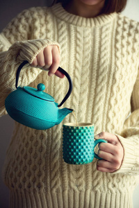 穿着保暖针织羊毛衫的妇女手持绿松石茶壶, 将凉茶倒入手工杯中。复制空间。冬季和圣诞节假期概念