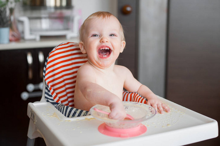 小可爱的女孩坐在高高的椅子和学习用勺子自己吃。有趣的快乐婴儿涂抹在桌子上的食物
