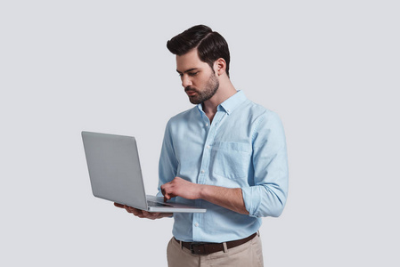 漂亮的年轻男子工作在他的笔记本电脑, 而站在反对灰色背景