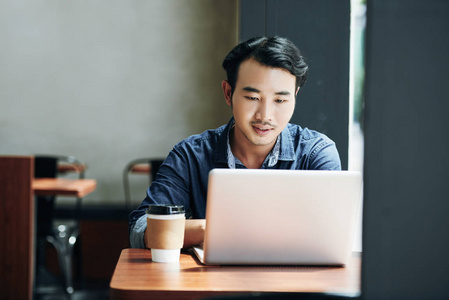 年轻的亚裔男子坐在咖啡表和工作在笔记本电脑