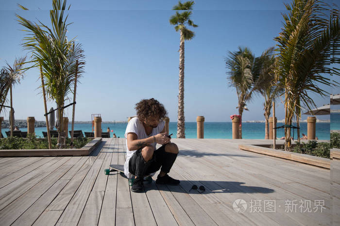 时尚男子坐在附近的海滩和使用智能手机
