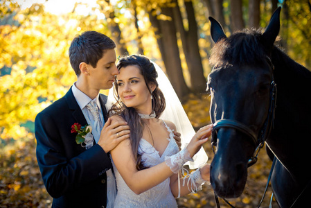 浪漫的肖像的时尚新郎温柔地拥抱他迷人的华丽的新娘在他们的步行与马在秋季公园