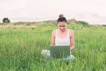 年轻美丽的亚洲妇女使用笔记本电脑在自然草地上